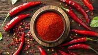 Ground red chili powder in wooden bowl surrounded by fresh hot peppers on rustic wooden background with scattered spices and bay leaves.