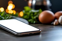 Modern smartphone with blank white screen on dark kitchen counter with fresh ingredients and warm bokeh lights in background, food photography setup.