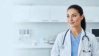 Young female doctor with stethoscope smiling confidently in modern medical office wearing white coat and blue scrubs for healthcare marketing.