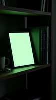 Tablet with glowing green screen displaying blank display on wooden shelf with books and headphones in modern dark interior setting for tech and lifestyle design projects.