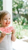 Toddler girl with blonde hair eating pink watermelon slice outdoors near window with green garden background, summer fruit enjoyment and childhood happiness.
