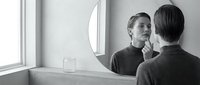 Young adult male with light skin tone and short hair examining face in bathroom mirror, touching chin in contemplative pose. Monochromatic setting with natural window light. Suitable for wellness, skincare, grooming, and personal care concepts.