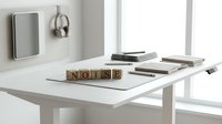 Minimalist white desk workspace with wooden letter blocks spelling NOISE, stacked books and modern wall-mounted speaker in bright natural light office interior.