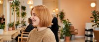 Young blonde woman smiling in modern cafe with warm lighting and plants. Casual lifestyle portrait for wellness, happiness, and social dining concepts.