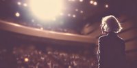 Silhouette of female speaker facing audience in theater auditorium with dramatic stage lighting and blurred crowd in background.