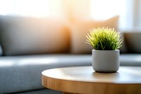 Decorative grass plant in modern gray ceramic pot on wooden round coffee table against blurred living room interior with soft window light.