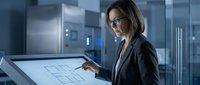 Middle-aged woman with brown hair and glasses wearing dark business blazer reviewing architectural blueprints on digital touchscreen display in modern office environment with glass partitions.