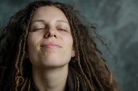 Young woman with curly hair and closed eyes expressing serenity and peace, smiling gently against dark background. Natural lighting highlights facial features.