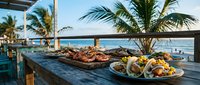 Grilled seafood platter with lobster and mixed dishes on wooden table at tropical beachfront restaurant during golden hour sunset with palm trees and ocean view.