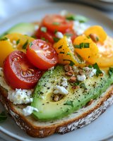 Fresh avocado toast topped with creamy feta cheese, colorful cherry tomatoes, pine nuts and herbs on rustic sourdough bread, served on ceramic plate.