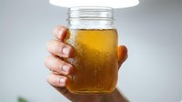 Hand holding glass jar filled with golden amber liquid, condensation droplets on exterior, isolated on white background, suitable for beverage, health supplement, honey, or organic product marketing and packaging design.