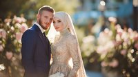 Young Muslim couple in wedding attire, groom in navy pinstripe suit and bride in gold embroidered dress with white hijab, posing against blurred floral background.