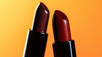Deep burgundy and metallic red lipsticks on vibrant orange background with dramatic lighting and reflective surfaces create luxurious cosmetic composition.