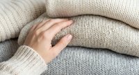 Cozy beige knitted sweaters stacked with woman's hand wearing ring touching soft wool fabric texture for autumn winter fashion and comfort clothing concepts.