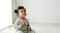 Toddler with dark curly hair and medium skin tone wearing sage green long-sleeve shirt, sitting on light bedding and looking upward with curious expression. Suitable for parenting, childcare, and family lifestyle content.