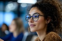 Young African American woman with curly hair and glasses smiling confidently in office environment, wearing brown sweater. Professional workplace portrait with soft bokeh background.