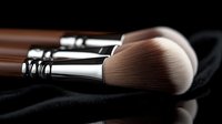 Professional makeup brushes with soft synthetic bristles and wooden handles on black reflective surface, close-up view showing luxury cosmetic tools detail.