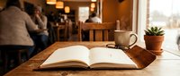 Open notebook with coffee cup and succulent plant on wooden table in cozy cafe with warm lighting and blurred customers in background for study workspace.