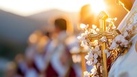 Golden ornate cross with delicate white flowers against blurred church ceremony background in warm sunset light, creating spiritual atmosphere for religious events.