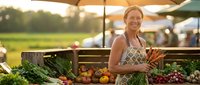Happy Caucasian woman vendor selling fresh organic vegetables at outdoor farmers market during golden hour sunset with abundant produce display.