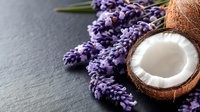 Fresh halved coconut with white flesh and purple lavender flowers arranged on dark stone background, natural spa and wellness composition.