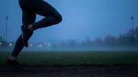 Runner jogging at dawn in misty blue hour atmosphere on grass field with city lights in background for fitness and wellness campaigns.
