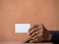 Senior male hand with dark skin tone holding blank white business card against warm beige background, suitable for corporate identity, business presentations, and professional marketing materials.