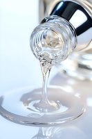 Clear viscous liquid pouring from glass bottle creating glossy stream and ripple effect on white surface, macro photography with soft focus and highlights.