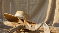 Woven straw sun hat with cream ribbon on driftwood against neutral linen backdrop. Natural summer fashion accessory for beach vacation and outdoor style.