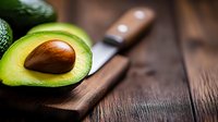 Fresh ripe avocado half with pit on rustic wooden cutting board with knife, healthy organic superfood ingredient for cooking and nutrition.