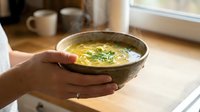 Hands holding ceramic bowl with creamy soup garnished with fresh herbs in bright kitchen setting for comfort food and homemade cooking concepts.