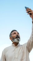 Mature man with gray beard and salt-and-pepper hair, South Asian ethnicity, wearing beige casual shirt, holding smartphone up against clear blue sky, looking upward with expression of wonder or searching for signal.