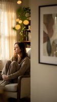 Young Caucasian woman in cozy sweater sitting thoughtfully in modern living room with warm lighting and plants for mental health and wellness concepts.