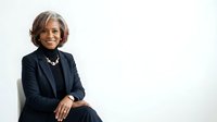 Senior woman with gray hair and warm smile wearing black blazer and pearl necklace, seated against white background for corporate portraits and professional headshots.