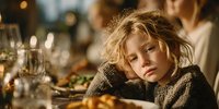 Child with tousled blonde hair resting head on hand at festive dinner table with bokeh lights and glassware in warm atmospheric setting.
