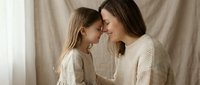 Young Caucasian mother and daughter sharing tender moment touching foreheads in cream sweaters against neutral fabric backdrop for family bonding concepts.