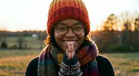 Young African American woman in colorful knit hat and scarf smiling with hands in prayer pose during golden hour sunset in rural field landscape.