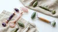 Assorted gemstone facial rollers with wooden and metal handles arranged on neutral linen fabric, featuring amethyst, rose quartz, and jade stones for skincare massage and wellness routines.
