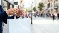Man in business attire holding blank white shopping bag on urban street with blurred pedestrians and storefronts in background. For retail, commerce, and shopping concepts.
