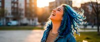 Young woman with blue hair laughing joyfully in urban park during golden hour sunset, expressing happiness and freedom in city environment.