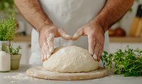 Male hands kneading fresh dough on wooden board with flour and herbs. Close-up view of homemade bread preparation in bright kitchen setting.