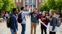 Diverse college students celebrating together on university campus courtyard with brick buildings and green trees in background during graduation or orientation day.