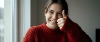 Young Asian woman in red knit sweater giving thumbs up gesture while smiling at camera near window. Positive attitude and approval concept for lifestyle content.