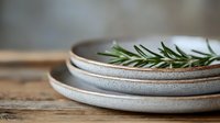 Fresh rosemary sprig on stacked ceramic plates with rustic texture and earthy tones, set against weathered wooden surface in minimalist composition.