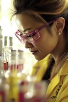 Asian female scientist in pink glasses examining laboratory samples with focused expression, wearing yellow lab coat against blurred research equipment background.