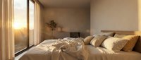 Cozy bedroom with unmade bed, golden bedding, decorative pillows, and warm sunlight streaming through large windows at sunset.