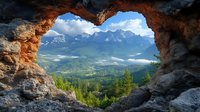 Natural rock arch frames dramatic mountain landscape with snow-capped peaks, green valley, and morning mist floating above pine forest in summer wilderness.