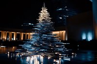 Modern illuminated spiral Christmas tree installation with glowing white lights creating geometric pattern, reflecting on wet surface against dark urban background.