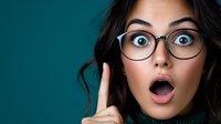 Young Asian woman with glasses showing surprised expression and raised finger against teal background, capturing genuine emotion and excitement in studio lighting.
