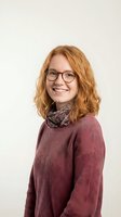 Happy young Caucasian woman with curly red hair and glasses wearing burgundy sweater and scarf smiling at camera on neutral background for lifestyle content.